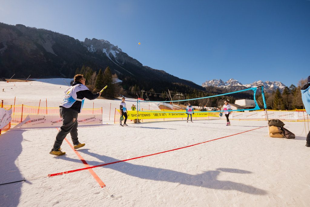 Campo da Snow Tennis tracciato sulla neve: rete azzurra, linee rosse sulla pista e giocatori in azione, con le montagne del Friuli alle spalle